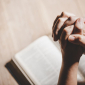 Auf einem Tisch liegt eine Bibel, &uuml;ber der Bibel sieht man H&auml;nde, die zum Gebet verschr&auml;nkt sind. 