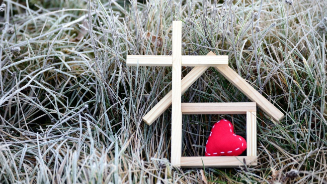 Symbolhafte Darstellung eines kleines Holzhauses, eines Holzkreuzes und eines roten Filzherzens auf gefrorener Wiese