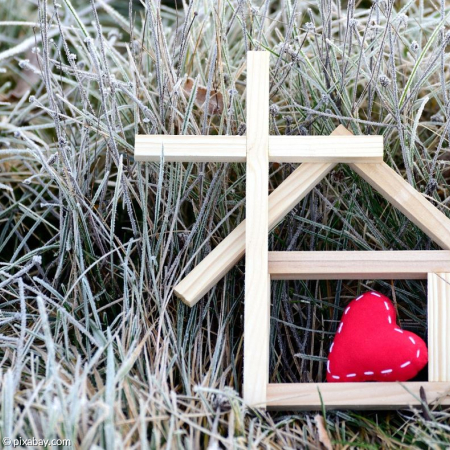 Symbolhafte Darstellung eines kleines Holzhauses, eines Holzkreuzes und eines roten Filzherzens auf gefrorener Wiese